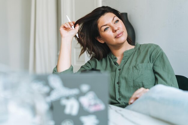 Młoda myśląca brunetka kobieta plus rozmiar w luźnej koszuli, pracująca przy laptopie w jasnym, nowoczesnym biurze