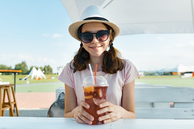 Młoda nastolatek dziewczyna w kapeluszowych okularach przeciwsłonecznych uśmiecha się chłodnego jagodowego koktajl i pije