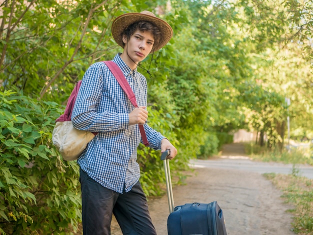 Młoda nastolatka chłopiec w letnim kapeluszu i przypadkowych ubraniach chodzi z walizką i stawia dalej plecaka