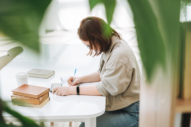 Młoda nastolatka studentka college'u w okularach odrabianiu lekcji z telefonem komórkowym w nowoczesnej bibliotece