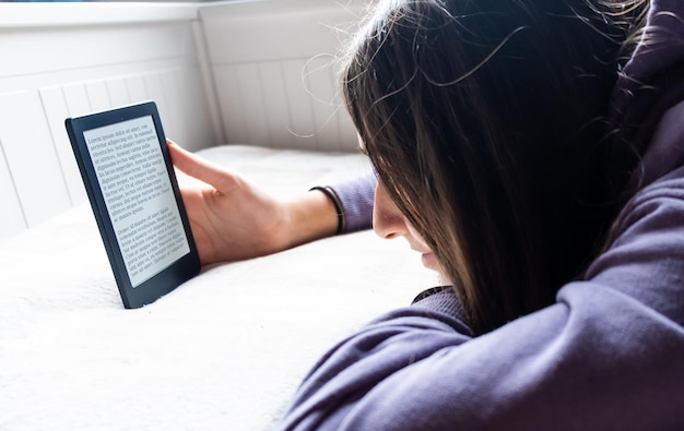 Zdjęcie młoda nastoletnia dziewczyna jest skupiona na czytaniu elektronicznego romansu ze swoim czytnikiem e-booków, gdy leży na łóżku. tekst na czytniku ebooków to „lorem ipsum”