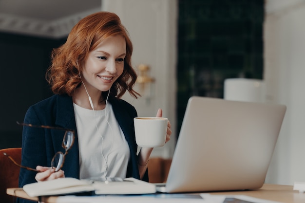 Młoda Nauczycielka Prowadzi Lekcję Online, Rozmawia Z Uczniami Za Pośrednictwem Laptopa, Organizuje Wideokonferencję, Uczy Uczniów, Pije Aromatyczną Kawę, Trzyma Szklanki W Dłoni, Ubrana Formalnie