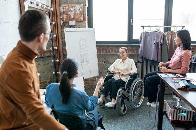 Młoda niepełnosprawna projektantka przy tablicy i innych współpracownikach omawia szkic nowego modelu z sezonowej kolekcji mody