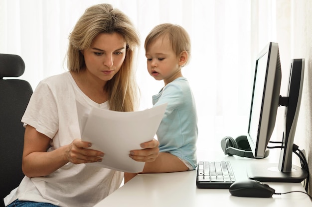 Zdjęcie młoda niezależna matka czytająca gazety w domowym biurze z synem siedzącym na biurku