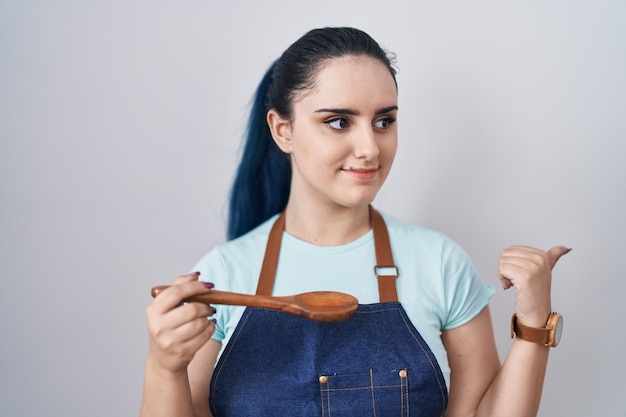 Młoda nowoczesna dziewczyna o niebieskich włosach ubrana w fartuch kucharza trzymająca łyżkę wskazująca kciukiem na bok uśmiechnięta zadowolona z otwartymi ustami