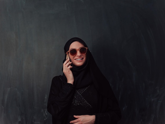 Młoda nowoczesna muzułmańska biznesmenka używająca smartfona w hidżabie przed czarną tablicą.