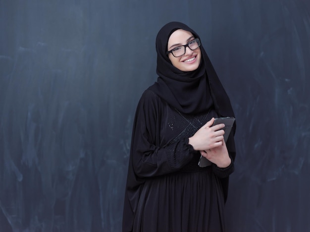 młoda, nowoczesna muzułmańska biznesowa kobieta używająca komputera typu tablet w okularach i ubraniach hidżabu przed czarną tablicą