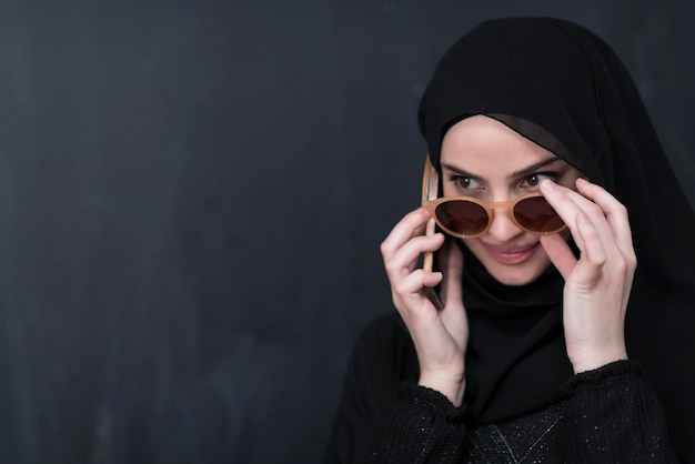 Młoda nowoczesna muzułmańska bizneswoman używająca smartfonów w okularach przeciwsłonecznych i hidżabie przed czarną tablicą.