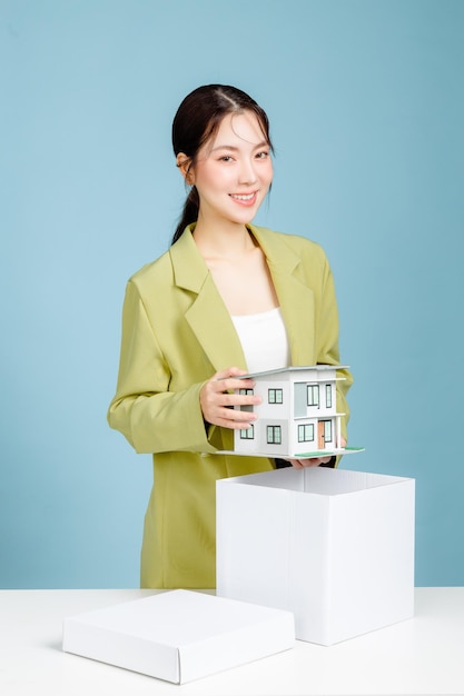 Młoda odnosząca sukcesy kobieta biznesu inwestora w swobodnym garniturze trzymając model domu przy białym biurku z laptopem pc odizolowanym na pastelowym niebieskim tle portret studyjny Koncepcja osiągnięcia