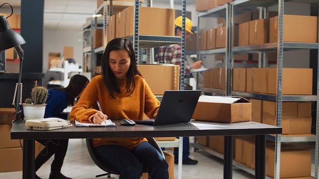 Młoda osoba robi inwentaryzację zapasów za pomocą laptopa, pisząc informacje logistyczne na dokumentach dotyczących rozwoju biznesu. Pracownica planująca dystrybucję produktów w przestrzeni magazynowej.