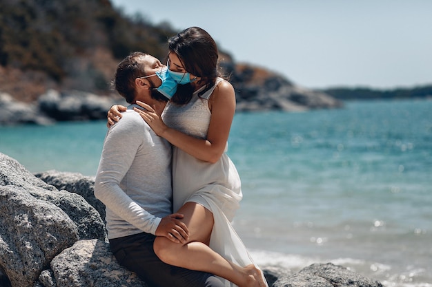 Młoda para całuje się seksualnie na plaży nad lazurowym morzem, nosząc maskę na twarz, która chroni ich przed infekcją z powodu wybuchu zakażenia koronowirusem COVID-19