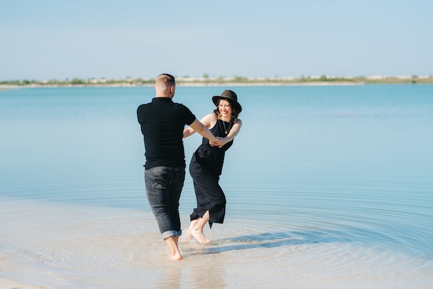 Młoda para facet z dziewczyną w czarnych ubraniach idą po białym piasku nad brzegiem błękitnej wody