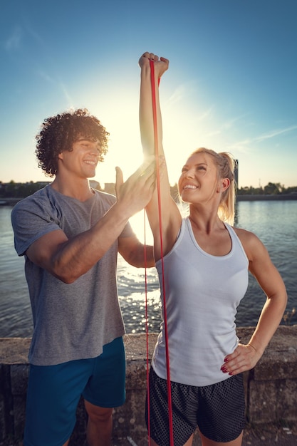 Młoda para fitness robi trening z gumką nad rzeką w zachód słońca. Kobieta wyciąga ramiona, a mężczyzna ją wspiera.