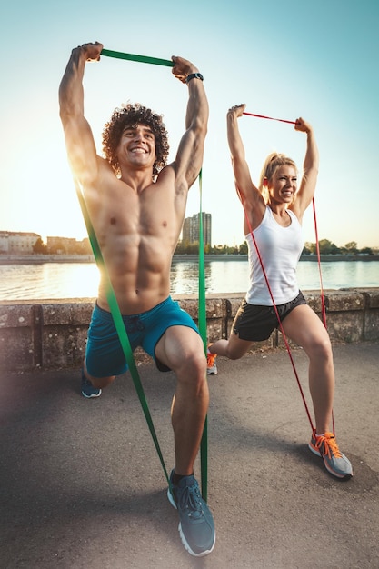 Młoda para fitness robi trening z gumką nad rzeką w zachód słońca.