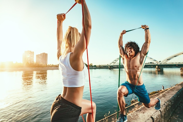 Młoda para fitness robi trening z gumką nad rzeką w zachód słońca.