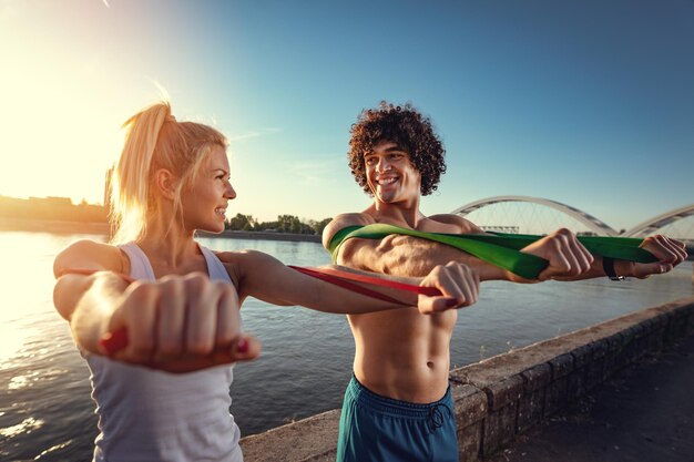 Młoda para fitness robi trening z gumką nad rzeką w zachód słońca.