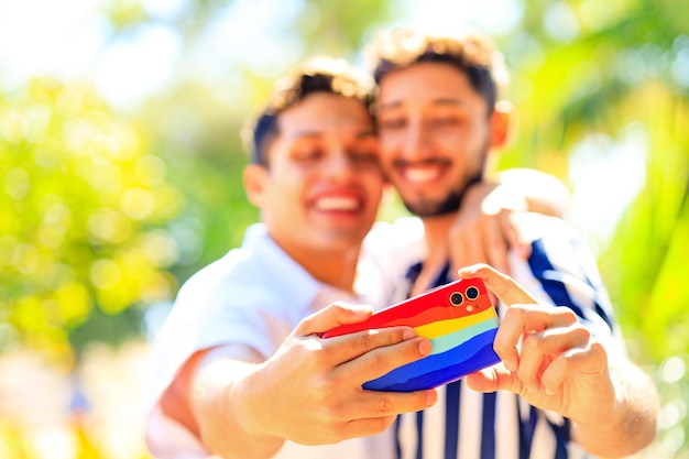 Młoda para gejów obejmująca i pokazująca swoją miłość wszystkim, którzy robią selfie na aparacie smartfona z tęczową obudową