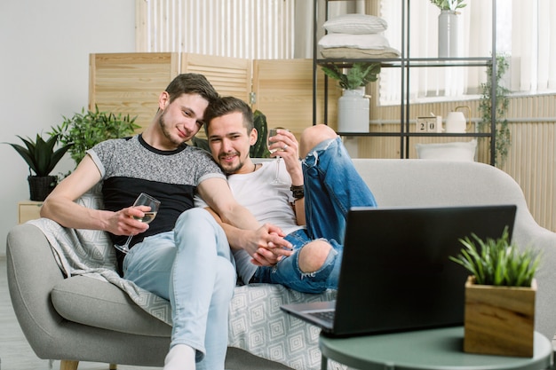 Młoda para gejów relaksujący wraz z winem na dużej wygodnej kanapie z laptopem, surfowanie po Internecie, widok z góry