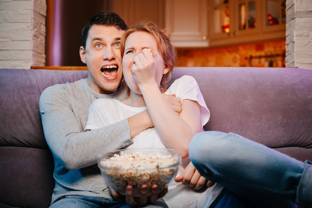 Młoda para jedząca popcorn i oglądająca film w domu na kanapie bardzo się boi