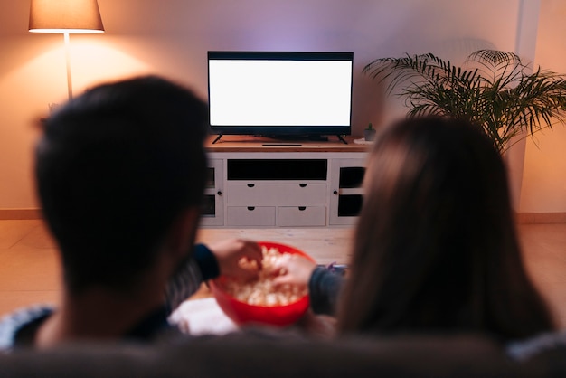 Młoda para kaukaski siedzi na kanapie, włączając telewizor, je popcorn.