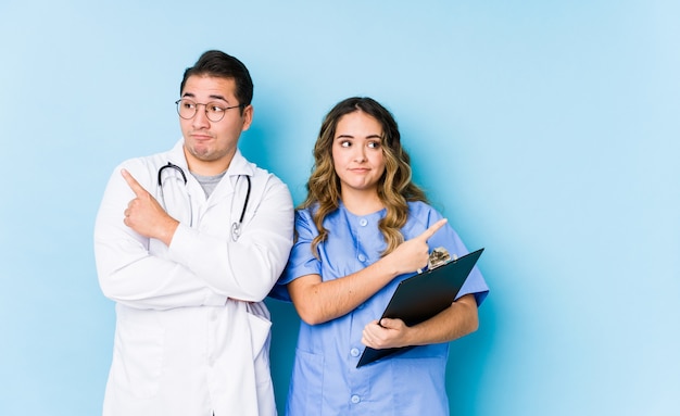 Młoda para lekarzy pozowanie w niebieskiej ścianie izolowanych punktów bokiem, próbuje wybrać jedną z dwóch opcji.