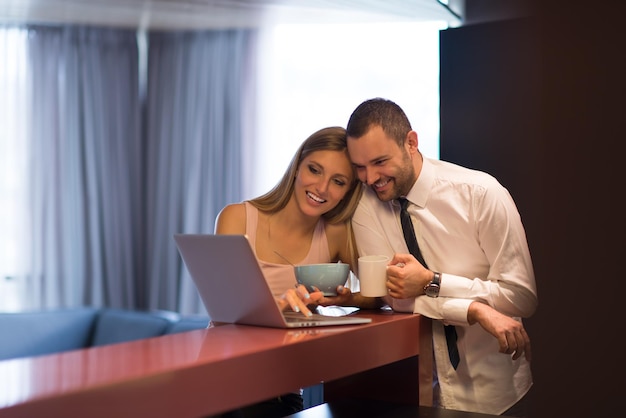 Młoda para przygotowuje się do pracy i korzysta z laptopa. Mężczyzna pije kawę, podczas gdy kobieta je razem śniadanie w luksusowym domu, patrząc na ekran, uśmiechając się.