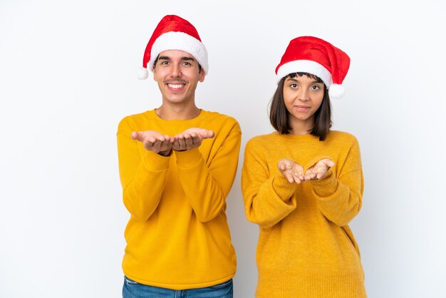 Młoda para rasy mieszanej świętuje Boże Narodzenie na białym tle trzymając wyimaginowaną copyspace na dłoni, aby wstawić reklamę