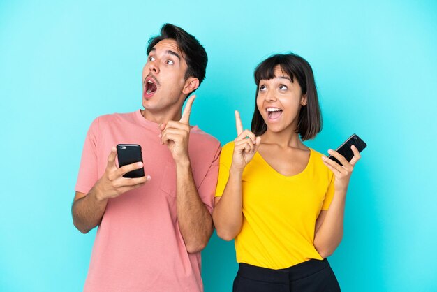 Młoda para rasy mieszanej trzymająca telefon komórkowy na białym tle na niebieskim tle, myśląca o pomyśle wskazującym palec w górę