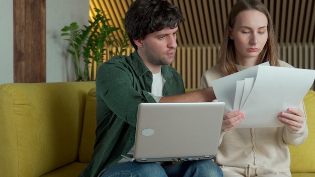 Młoda para siedzi na żółtej kanapie w domu i zarządza budżetem za pomocą laptopa