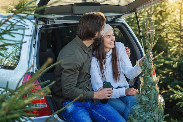 Młoda para siedzi w bagażniku swojego SUV-a, trzymając świeżo ściętą choinkę na plantacji