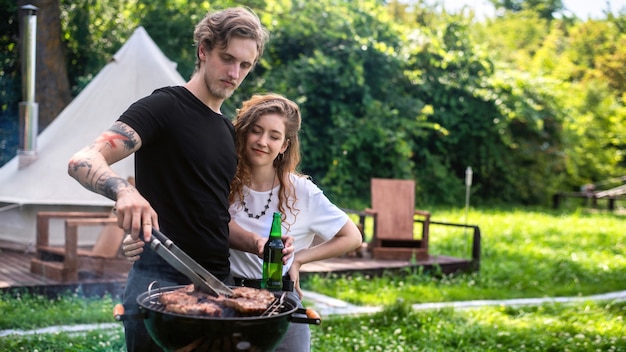 Młoda Para Smażenia Mięsa Z Grilla I Picia Piwa. Wokół Zieleń. Glamping