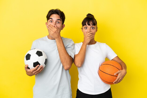 Młoda para sportowa grająca w piłkę nożną i koszykówkę na żółtym tle zakrywająca usta rękami za powiedzenie czegoś niewłaściwego