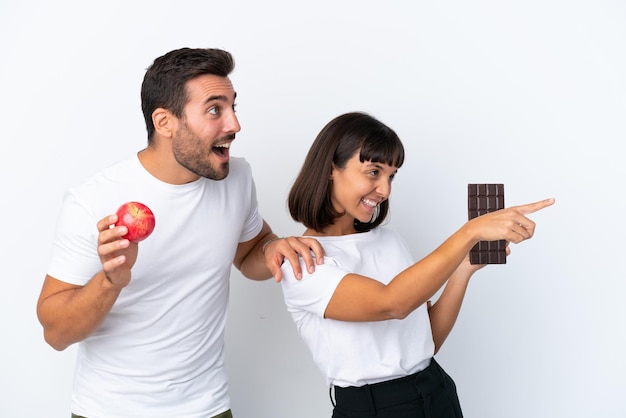 Młoda para trzymająca czekoladę i jabłko na białym tle wskazując na bok, aby zaprezentować produkt