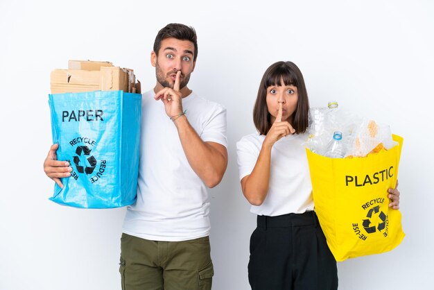 Młoda para trzymająca torbę pełną plastiku i papieru do recyklingu na białym tle na białym tle pokazując znak ciszy gest wkładania palca do ust