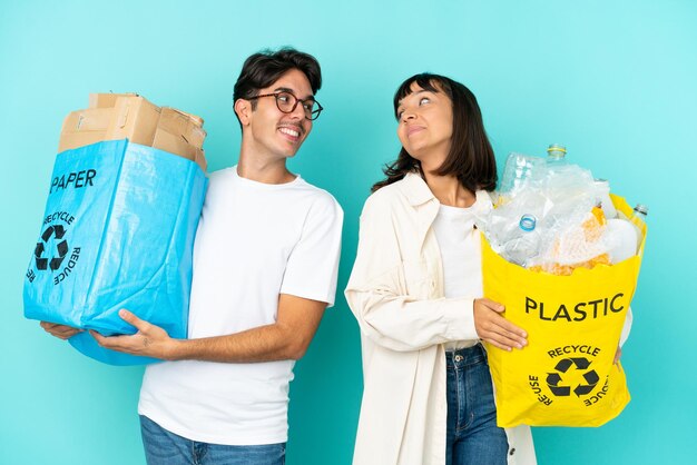 Młoda para trzymająca torbę pełną plastiku i papieru do recyklingu, na białym tle na niebieskim tle, patrząc przez ramię z uśmiechem