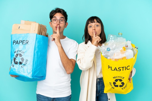 Młoda para trzymająca torbę pełną plastiku i papieru do recyklingu na białym tle na niebieskim tle pokazując znak ciszy gest wkładania palca do ust