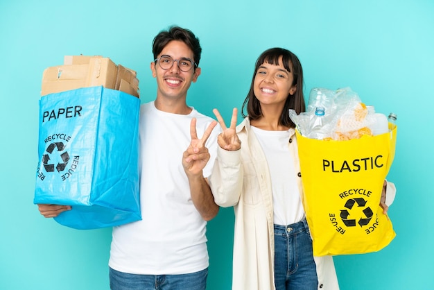 Młoda para trzymająca torbę pełną plastiku i papieru do recyklingu, na białym tle na niebieskim tle, uśmiechając się i pokazując znak zwycięstwa