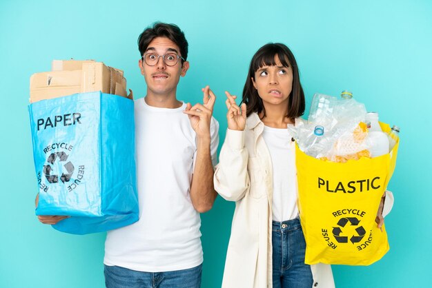 Młoda para trzymająca torbę pełną plastiku i papieru do recyklingu na białym tle na niebieskim tle z palcami skrzyżowanymi i życzącymi wszystkiego najlepszego