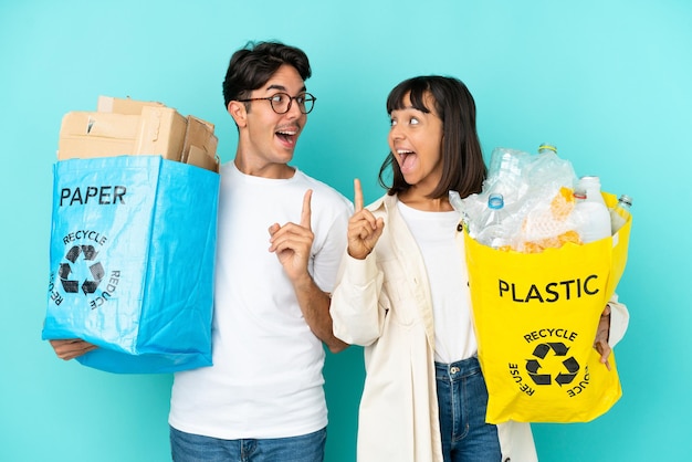 Młoda para trzymająca torbę pełną plastiku i papieru do recyklingu na białym tle na niebieskim tle, zamierzająca zrealizować rozwiązanie, podnosząc palec w górę