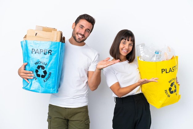 Młoda para trzymająca torbę pełną plastiku i papieru do recyklingu na białym tle wskazującą tyłem i prezentującą produkt