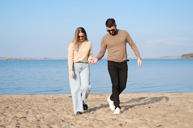 Młoda para w dżinsach i swetrach trzymając się za ręce i spacerując po plaży w słoneczną pogodę