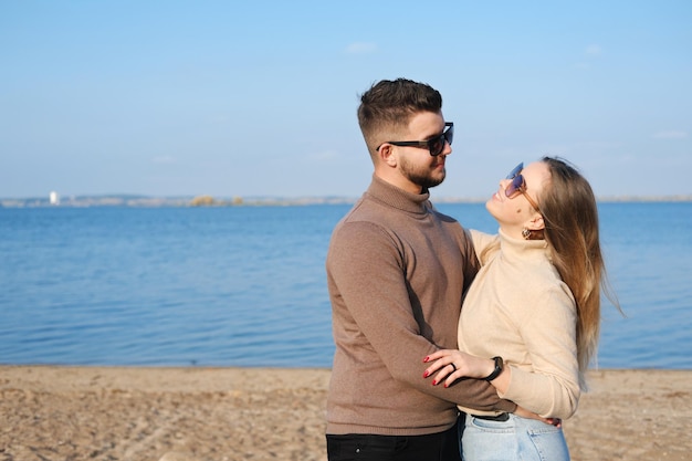 Młoda Para W Dżinsowych Swetrach I Okularach Przeciwsłonecznych Stoi Na Plaży W Pobliżu Jeziora I Przytula Się