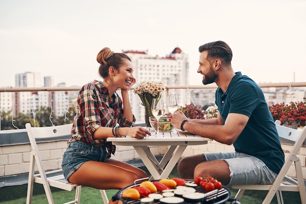 Młoda para w swobodnym ubraniu delektuje się romantyczną kolacją i uśmiecha się, delektując romantyczną kolację na tarasie na dachu na świeżym powietrzu