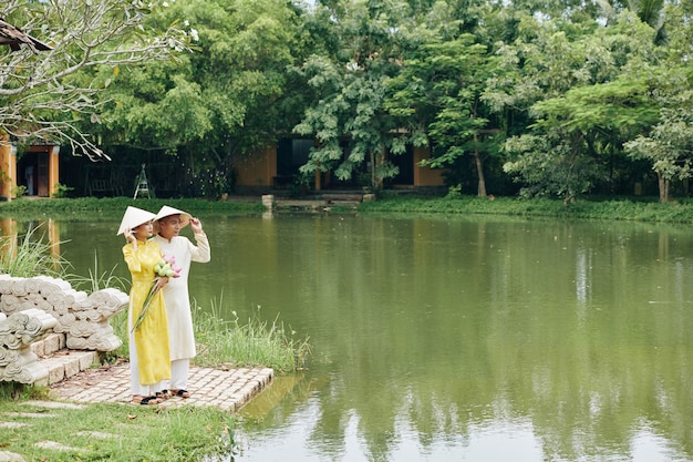 Młoda para wietnamska w sukniach ao dai i stożkowatych kapeluszach stoi po ślubie nad małym stawem
