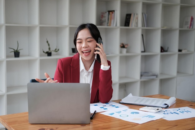 Młoda, pewna siebie azjatycka bizneswoman w czerwonym garniturze rozmawia telefonicznie ze swoim klientem, siedząc w swoim biurze.