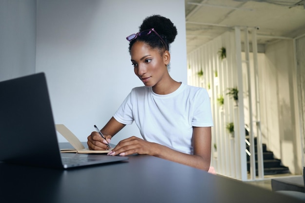 Młoda, pewna siebie, swobodna, afroamerykańska kobieta pracująca na laptopie i pisząca notatki w nowoczesnej przestrzeni co-workingowej