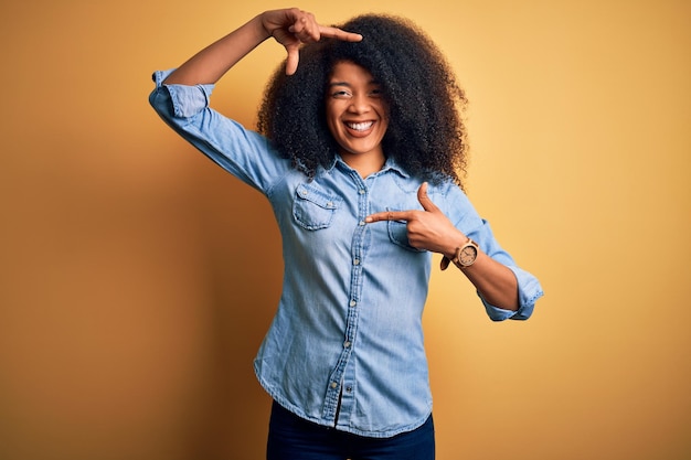 Młoda piękna Afroamerykanka z włosami afro stojąca nad żółtym odizolowanym tłem uśmiechnięta robiąca ramę dłońmi i palcami z radosną twarzą Kreatywność i koncepcja fotografii