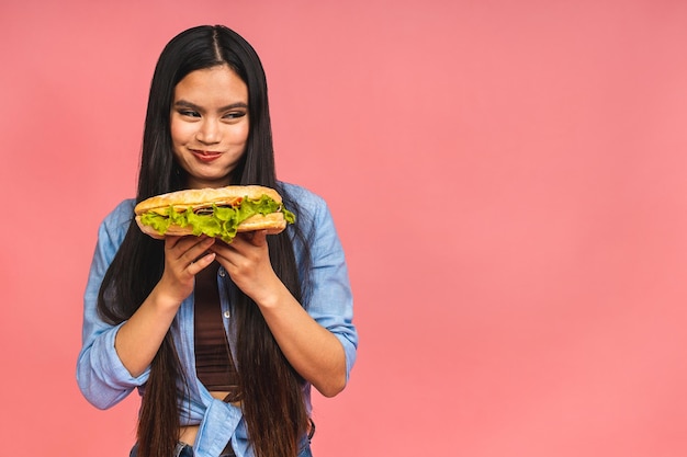 Młoda piękna azjatycka japońska chińska kobieta je kanapkę lub duży burger z satysfakcją Dziewczyna cieszy się smacznym hamburgerem na wynos koncepcja diety stojąc na białym tle nad różowym tle