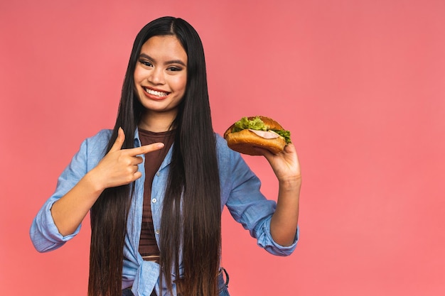 Młoda piękna azjatycka japońska chińska kobieta je kanapkę lub duży burger z satysfakcją Dziewczyna cieszy się smacznym hamburgerem na wynos koncepcja diety stojąc na białym tle nad różowym tle