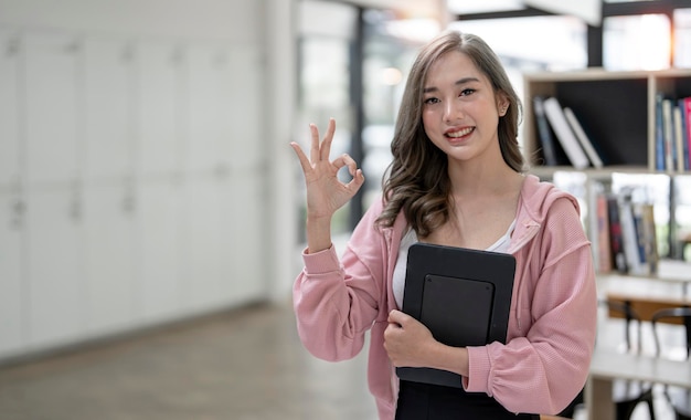 Młoda piękna azjatycka kobieta uśmiecha się i pokazuje znak OK na ręce stojąc w biurze z miejscem na kopię Patrząc na kamerę